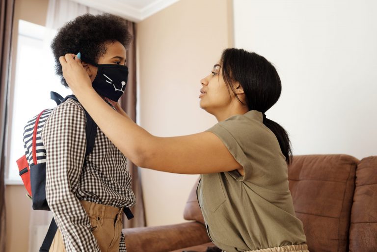 Mother putting a mask on her child, who is also wearing a backpack. Photo by August de Richelieu from Pexels