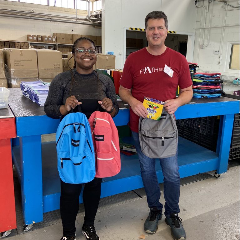 Two volunteers smile while holding backpacks full of school supplies