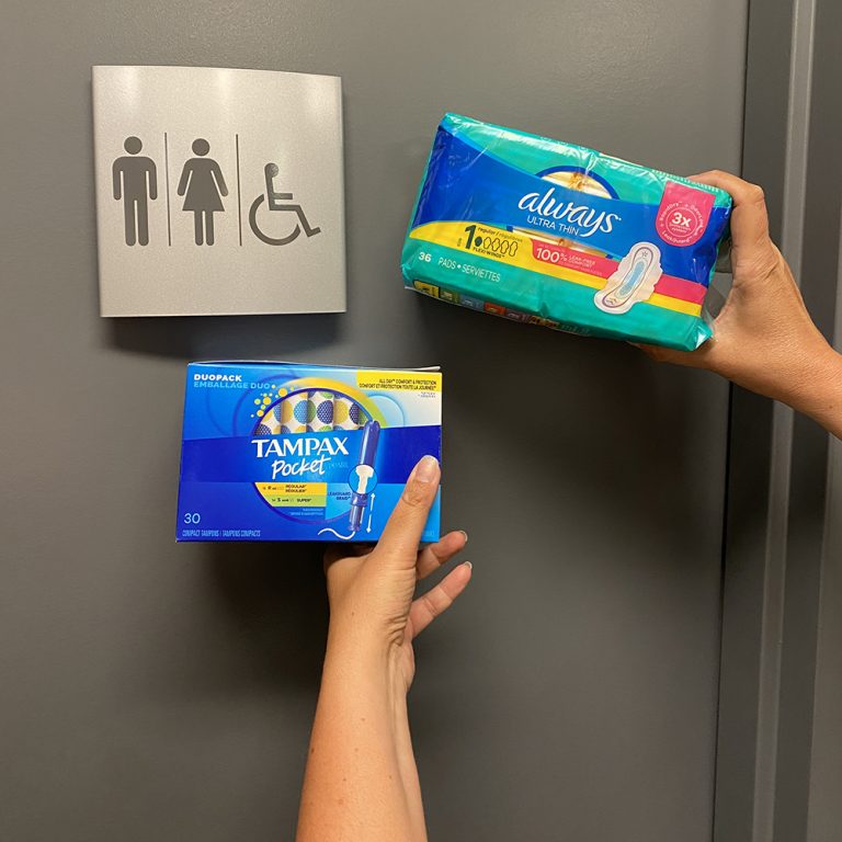 Two hands hold period products up outside of an all-gender washroom sign