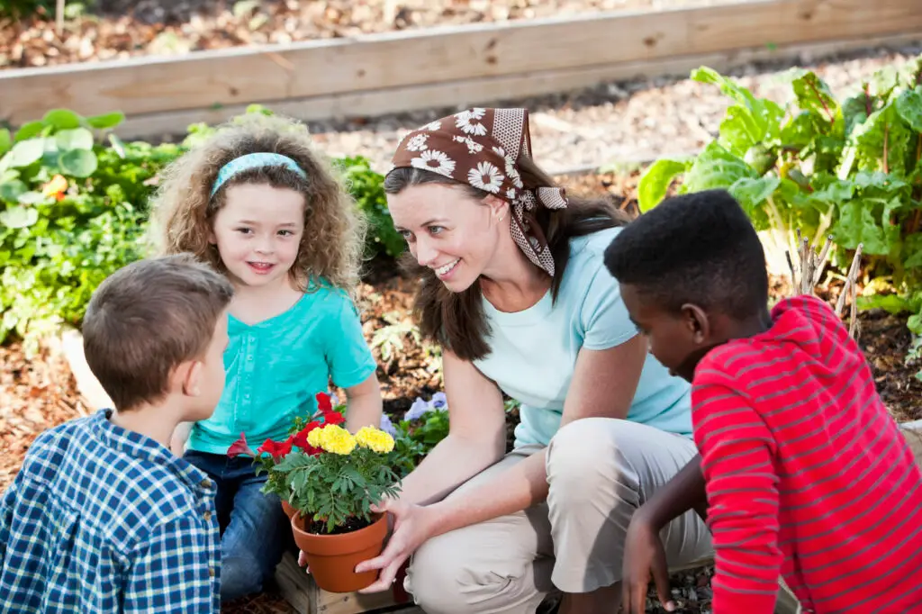 Woman (30s) with children (5, 6 and 10 years) planting flowers in community garden. Vegetables growing in planter. Focus on woman.