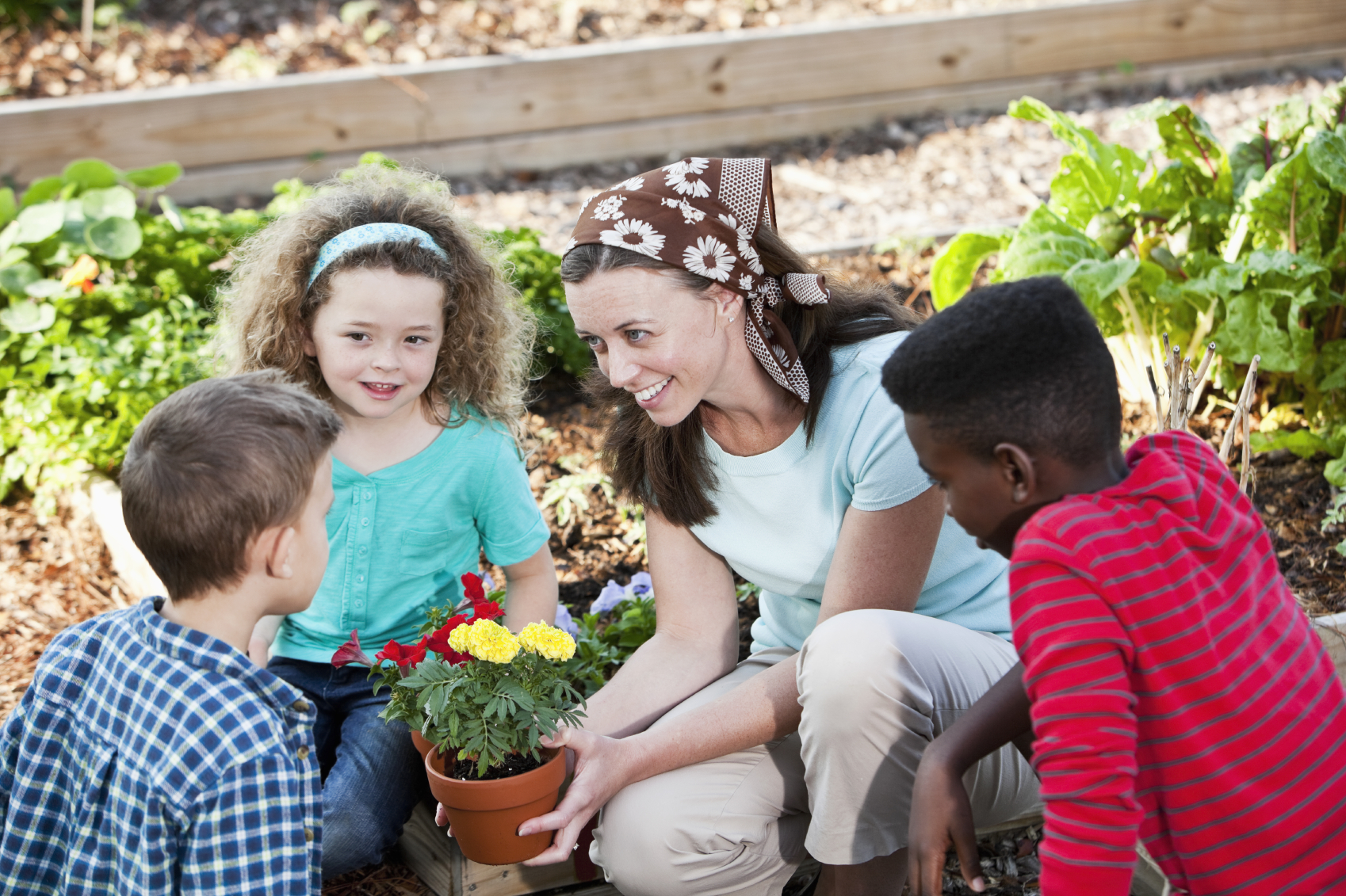 Woman (30s) with children (5, 6 and 10 years) planting flowers in community garden. Vegetables growing in planter. Focus on woman.