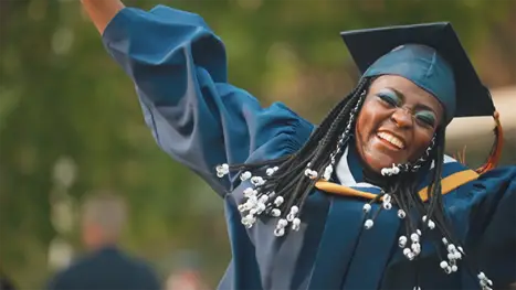 A young woman wearing a cap and gown celebrates her graduation outside a school.