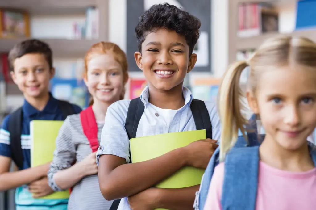 Portrait of happy multiethnic children holding books and wearing backpack at primary school.