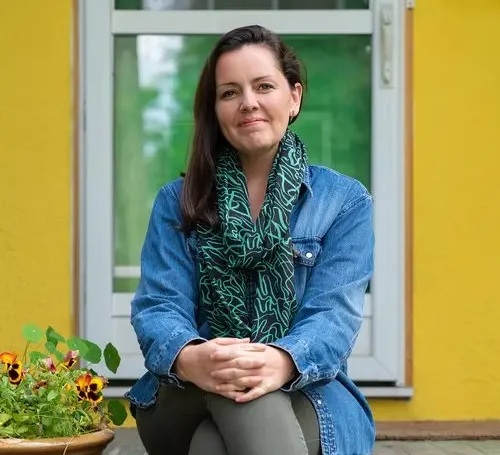 Artist Amanda Schutz poses with legs crossed on the front stoop of a yellow house.