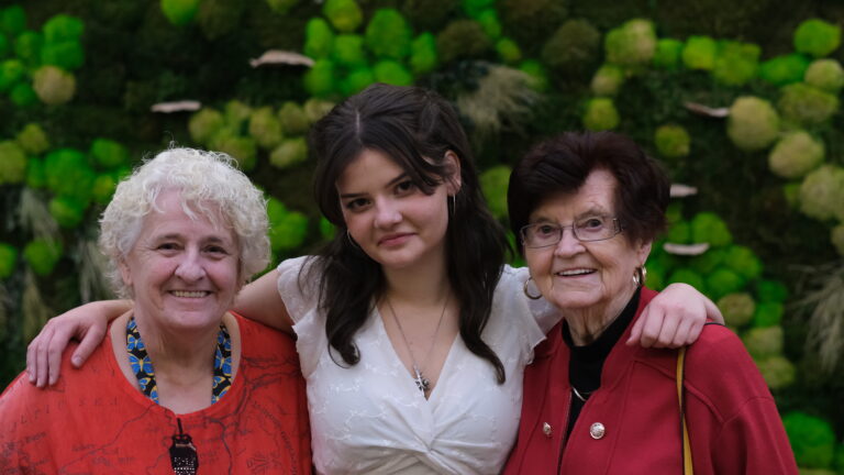 A teenaged girl stands between two smiling senior women in front of a living plant wall.