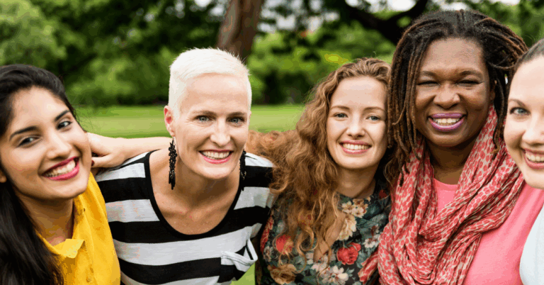 Group of 5 smiling women of various ethnicities and ages stand with their arms across each others shoulders.