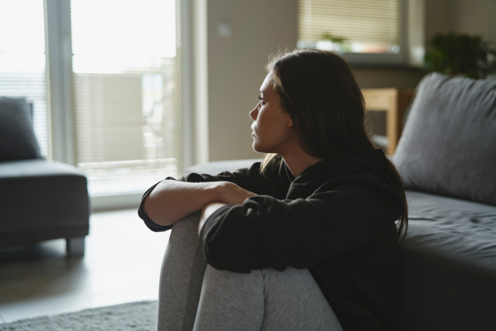Sad woman sitting on the floor and looking through window at home